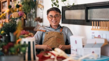 Flower shop owner wearing glasses and an apron sits behind a worktable surrounded by flowers, packaging supplies, and small boxes.