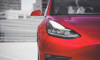 A close up of the right front of a red Tesla in front of a blurred cityscape background.