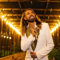 A man with long blonde hair, wearing a white tuxedo jacket, looks upwards.