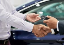 Two professionals shake hands while exchanging a car key fob beside a vehicle, symbolizing a vehicle sale, lease agreement, or dealership transaction.