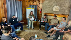 Group of men and women seated in a circle around a room as one woman stands and leads discussion.