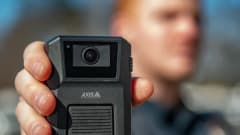 Close-up of a person holding an Axis body-worn camera toward the viewer. The camera is in sharp focus, while the person wearing a dark uniform appears blurred in the background outdoors.