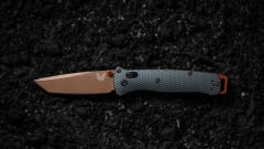 Benchmade Bailout folding knife with a bronze tanto blade, taiga green handle, and orange accents displayed open against a textured black background, highlighting its lightweight tactical design.