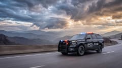 Ford Expedition police responder SUV drives along a winding mountain highway with red and blue emergency lights flashing beneath a dramatic cloudy sky.