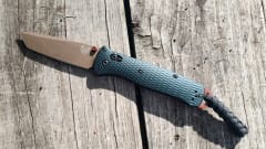 Open Benchmade folding knife with a bronze-colored blade, textured dark handle, and braided lanyard resting on weathered wooden boards.