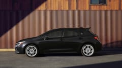 Side profile of a black 2027 Toyota Corolla Hatchback parked in front of a wood-paneled building, showing the compact hatchback's available 18-inch gloss white alloy wheels and updated styling.