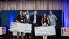 Group of five people on stage holding large checks at a Reynolds and Reynolds event, recognizing winners of an automotive innovation contest.