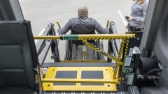 A wheelchair user operates a school bus wheelchair lift equipped with BraunAbility’s LiftSentinel barrier system as another person stands nearby with a handheld control.