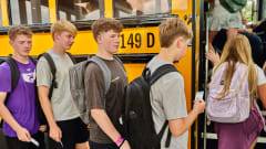 Students wearing backpacks line up to board a yellow school bus, each holding an NFC-enabled student ID card to scan as they enter. The image illustrates student ridership tracking technology that records boarding activity in real time.