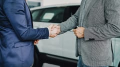 Photo of two men in suit jackets shaking hands next to new car inside of a dealership