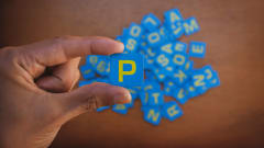 Photo of man's hand holding a square blue piece with a yellow capital P on it, with other letter pieces below on a surface