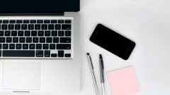 Photo of laptop on desk with pens, phone and sticky notepad beside it