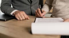 Photo of a man signing a paper at a desk while a man in a suit looks on 