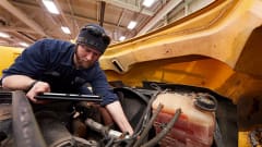A mechanic in a workshop leans over the open engine compartment of a large yellow vehicle, inspecting components while holding a tablet.