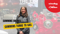 Andrea Lukas of Cummins stands beside a large red engine display at ACT Expo, with on-screen text reading “Cummins Turns to Gas” during a School Bus Fleet interview about new gasoline engine technology.