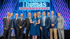 Group of Thomas Built Buses executives, employees, and award recipients pose onstage during an awards ceremony beneath a large Thomas Built Buses sign, with one woman holding a glass trophy at center stage.