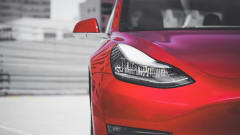 A close up of the right front of a red Tesla in front of a blurred cityscape background.