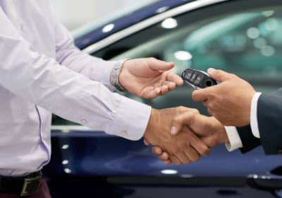 Two professionals shake hands while exchanging a car key fob beside a vehicle, symbolizing a vehicle sale, lease agreement, or dealership transaction.