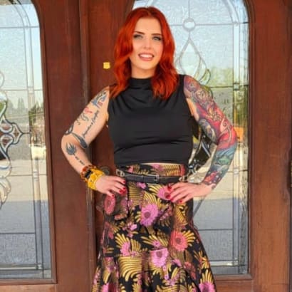 A woman with red hair and her arms akimbo, standing in front of glass doors. 