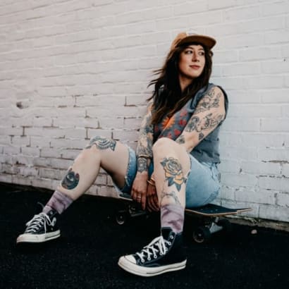 A young woman in jean shorts, wearing a baseball hat, leans against a brick wall.