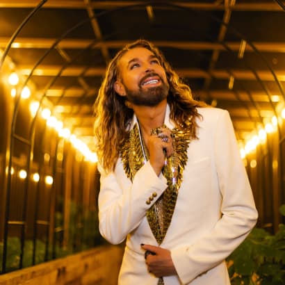 A man with long blonde hair, wearing a white tuxedo jacket, looks upwards. 