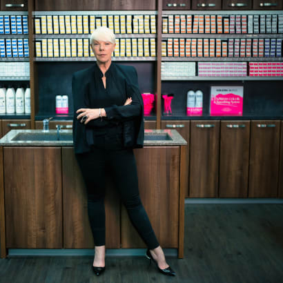 A woman in black and with short blonde hair, with arms crossed, standing in front of shelves of beauty products.