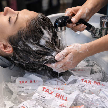 A woman get her hair rinsed at a shampoo bowl in a salon, but instead of suds in the bowl, there are tax bills.