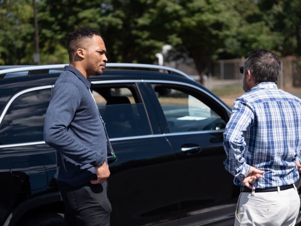 Two men converse next to a vehicle in ride and drive. 
