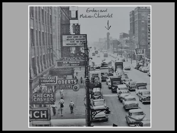 Black-and-white photo of a busy city street in the 1950s with vintage cars, storefront signs, pedestrians, and handwritten notes pointing to the distant Emkay and Midway Chevrolet location.