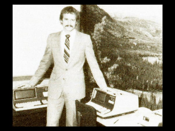 Vintage sepia-toned photo of a man in a light-colored suit standing beside early office computer equipment.