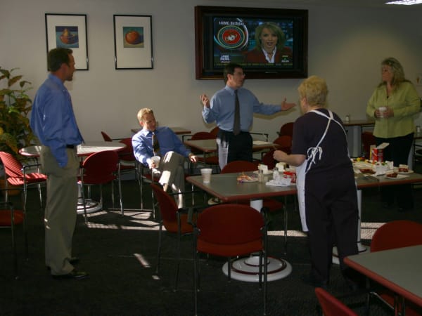 Employees gathered in an office break room during a workplace celebration or meeting, with refreshments on tables and a television playing in the background.