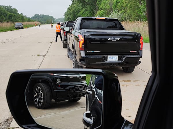 View out driver's window looking over the side-view mirror at other black pickup trucks waiting in line ahead.