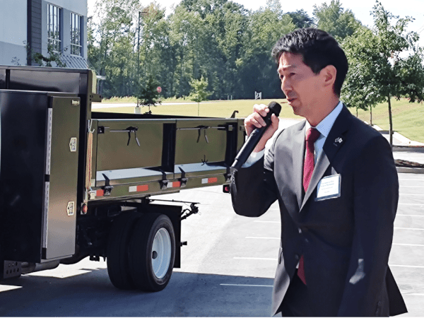 A business man holds a microphone and speaks with a truck in the background.