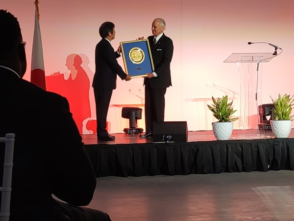 Two men in business suit exchange gifts on a stage.