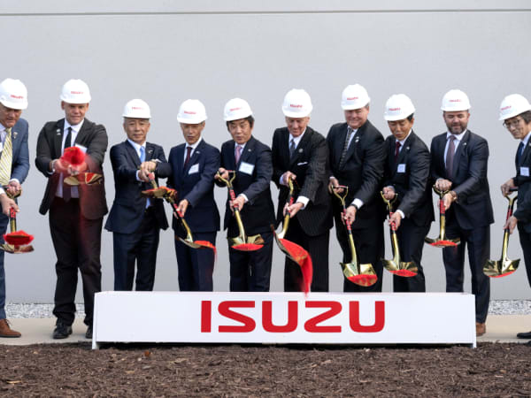 Men in suits and hardhats shovel red dirt in front of a box with Isuzu logo.