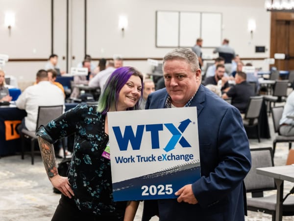 Lauren Fletcher and Wayne Parham posing with an attendee holding a Work Truck Exchange 2025 sign inside the meeting room filled with one-on-one business meetings.