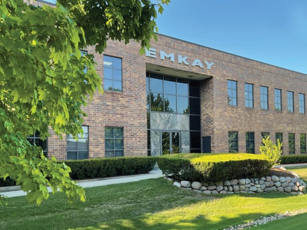 Modern brick office building with large EMKAY signage, surrounded by trees, landscaping, and a manicured lawn on a sunny day.