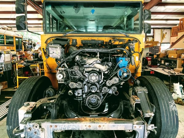 A yellow school bus sits inside a maintenance shop with its front bodywork removed, exposing the engine, belts, pulleys, and mechanical components. The image offers a detailed look at a bus undergoing major repair or maintenance.