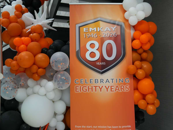 EMKAY 80th anniversary display featuring an orange commemorative banner surrounded by black, white, and orange balloon decorations.