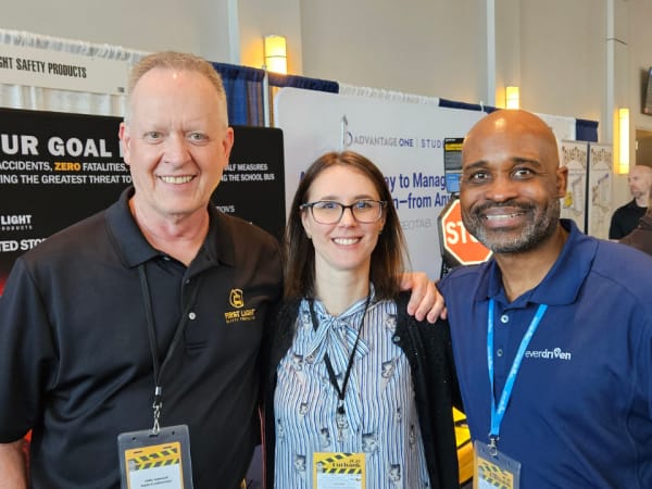 Three people smile together on the vendor expo floor, standing in front of exhibit booths and safety product displays.