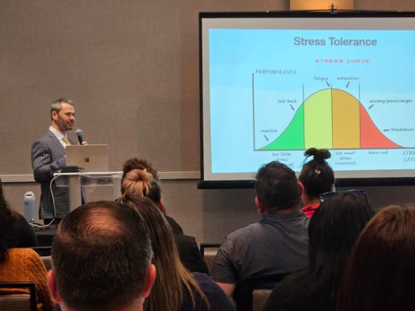 A speaker presents on stress tolerance and burnout, standing beside a projected graph illustrating performance levels and stress curves during an educational session.