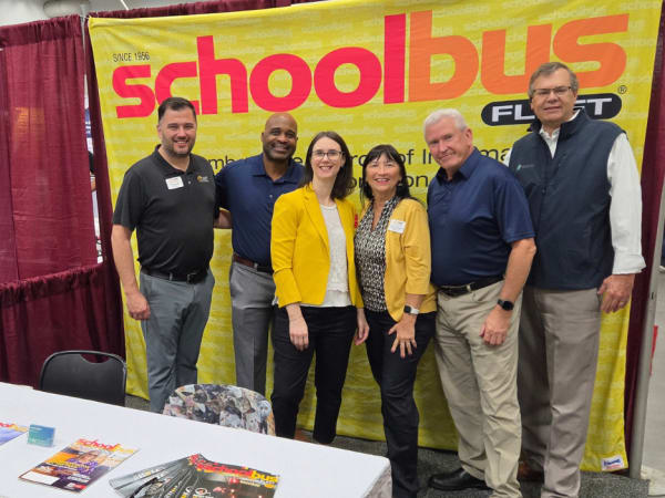 Six school transportation industry professionals pose together in front of the School Bus Fleet banner at the NAPT ACTS 2026 trade show.