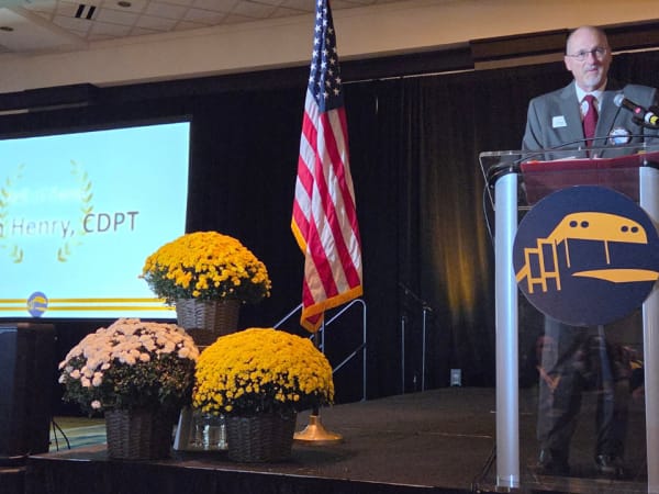 A speaker addresses attendees from the NAPT ACTS 2026 podium as a screen beside the stage displays the name of Hall of Fame honoree Keith Henry, CDPT.
