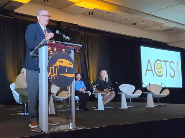 A speaker addresses attendees from the NAPT ACTS 2026 podium, with other presenters seated behind him and the conference logo displayed on a large screen.