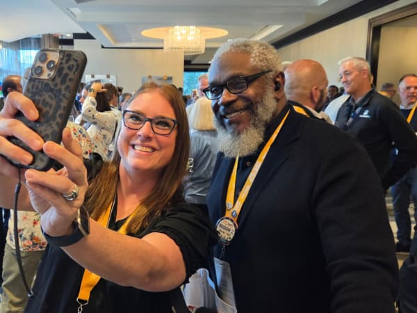 Two NAPT ACTS 2026 attendees smile for a selfie during a busy conference networking event, with other school transportation professionals gathered behind them.
