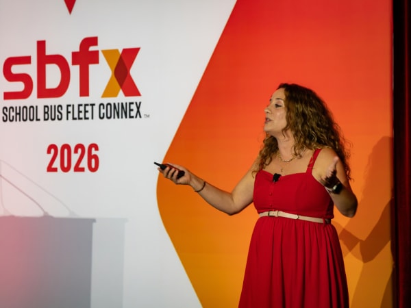 A presenter gestures while speaking onstage in front of the 2026 School Bus Fleet Connex backdrop.