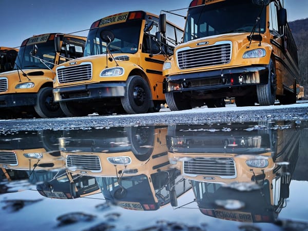 Several parked yellow school buses are reflected in a large puddle on a cold morning, creating a mirror image of the buses and blue sky across the water.