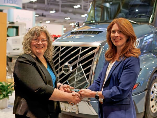 Two women shaking hands with Top 20 trophy between them and Volvo truck in background
