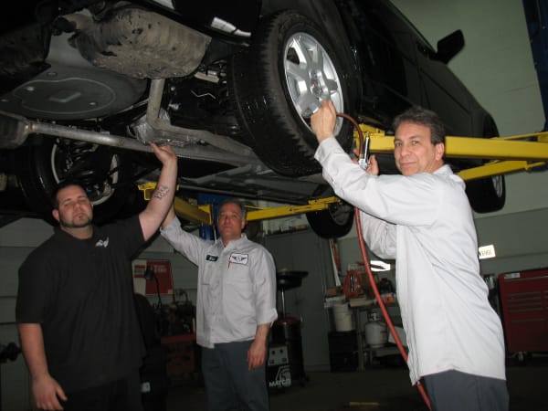 Three automotive technicians working beneath a vehicle raised on a service lift in a repair garage.