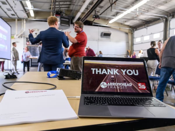 Closing slide on a laptop from the Arrowhead Transit RideSmart 2025 launch event as the crowd mingles in the background.